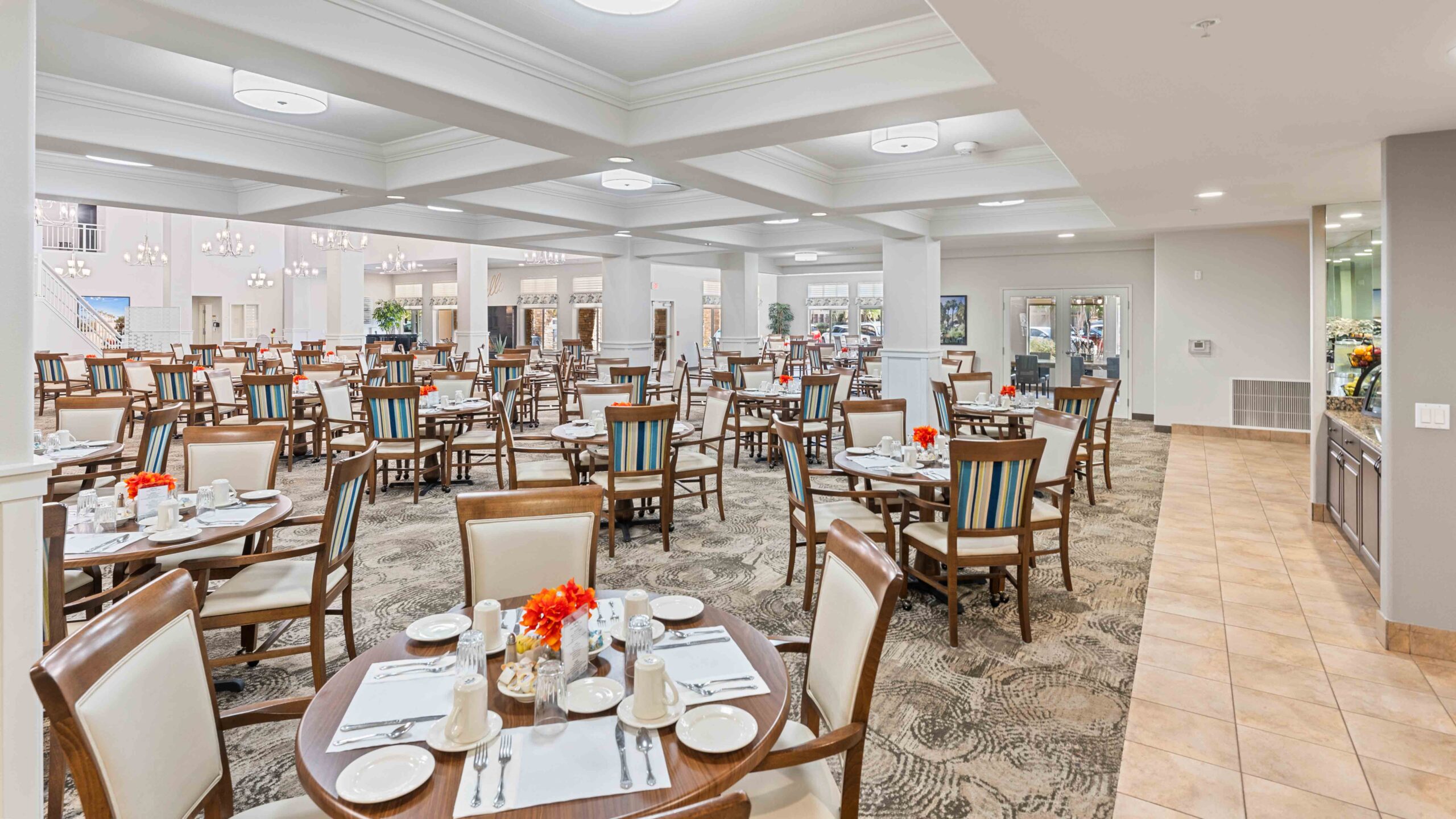 A spacious, elegant dining hall with neatly set wooden tables and chairs, bright lighting, floral centerpieces, and large windows letting in natural light. The room has a clean, welcoming atmosphere.