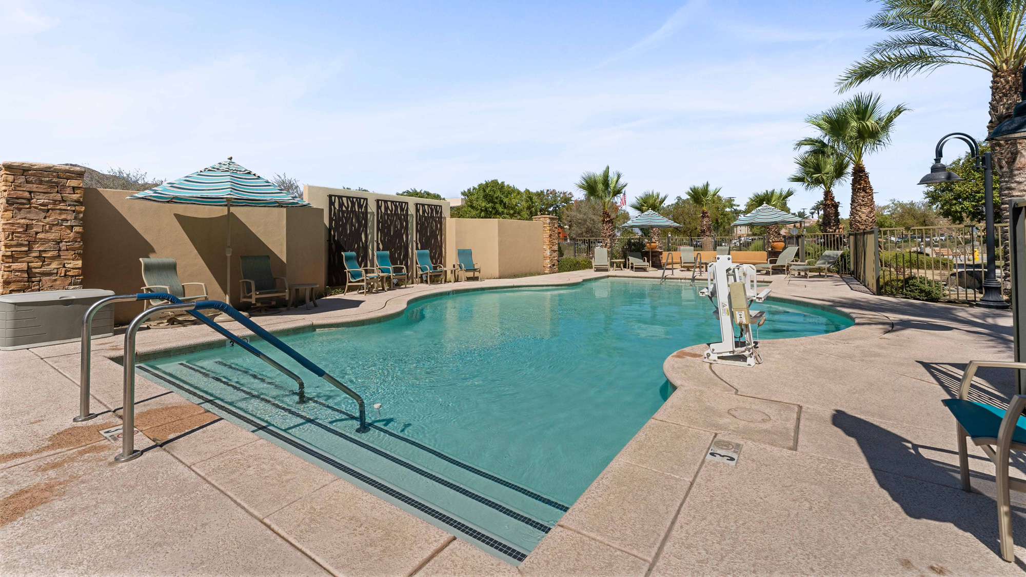 Outdoor swimming pool with clear water, poolside chairs, loungers, umbrellas, palm trees, and a stone wall under a blue sky; accessible pool lift and handrails are visible by the pool steps.