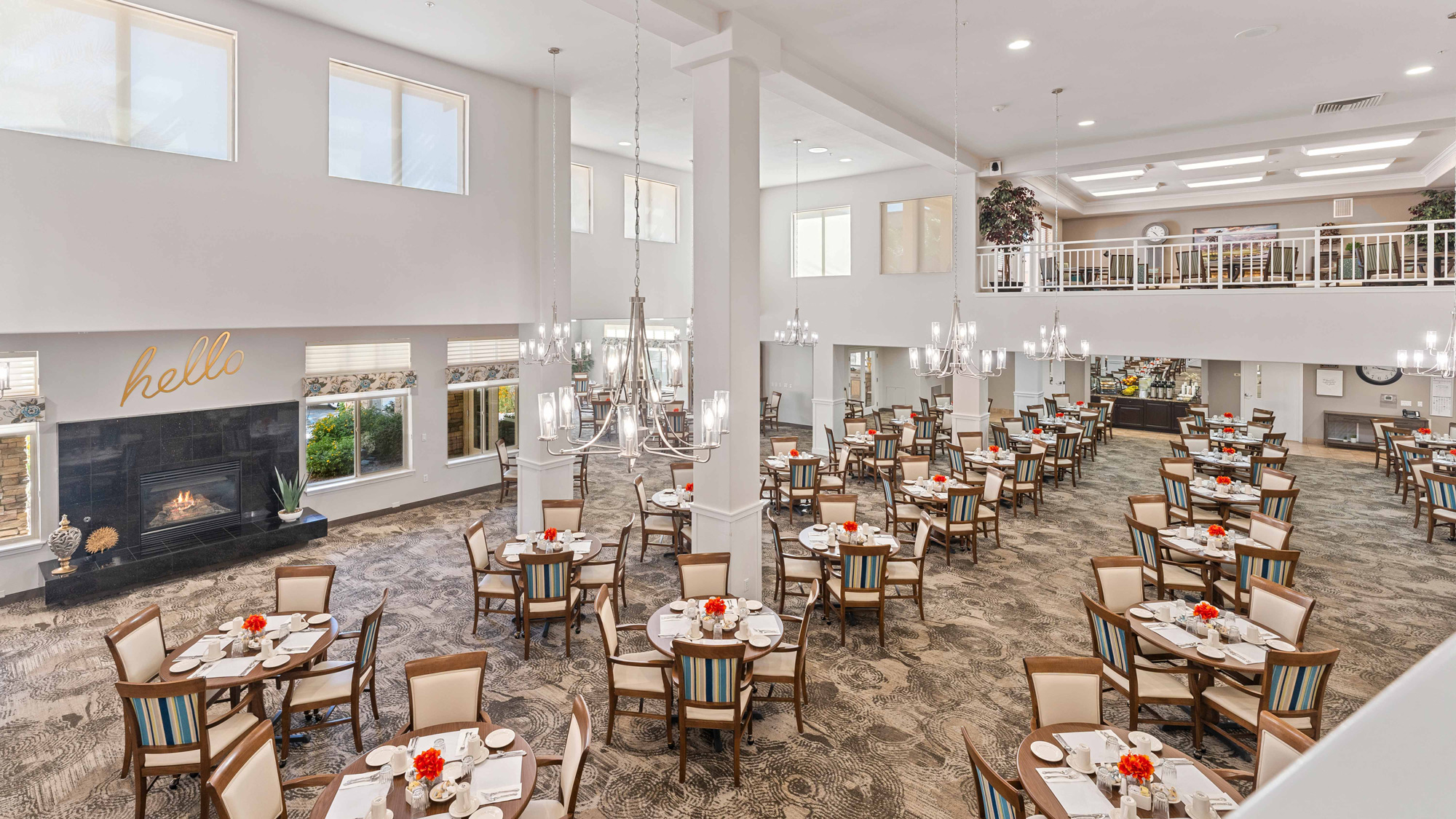 A spacious, elegantly decorated dining room with many round tables, each set with white dishes and red flower centerpieces. Large windows, chandeliers, and a fireplace create a bright, welcoming atmosphere.