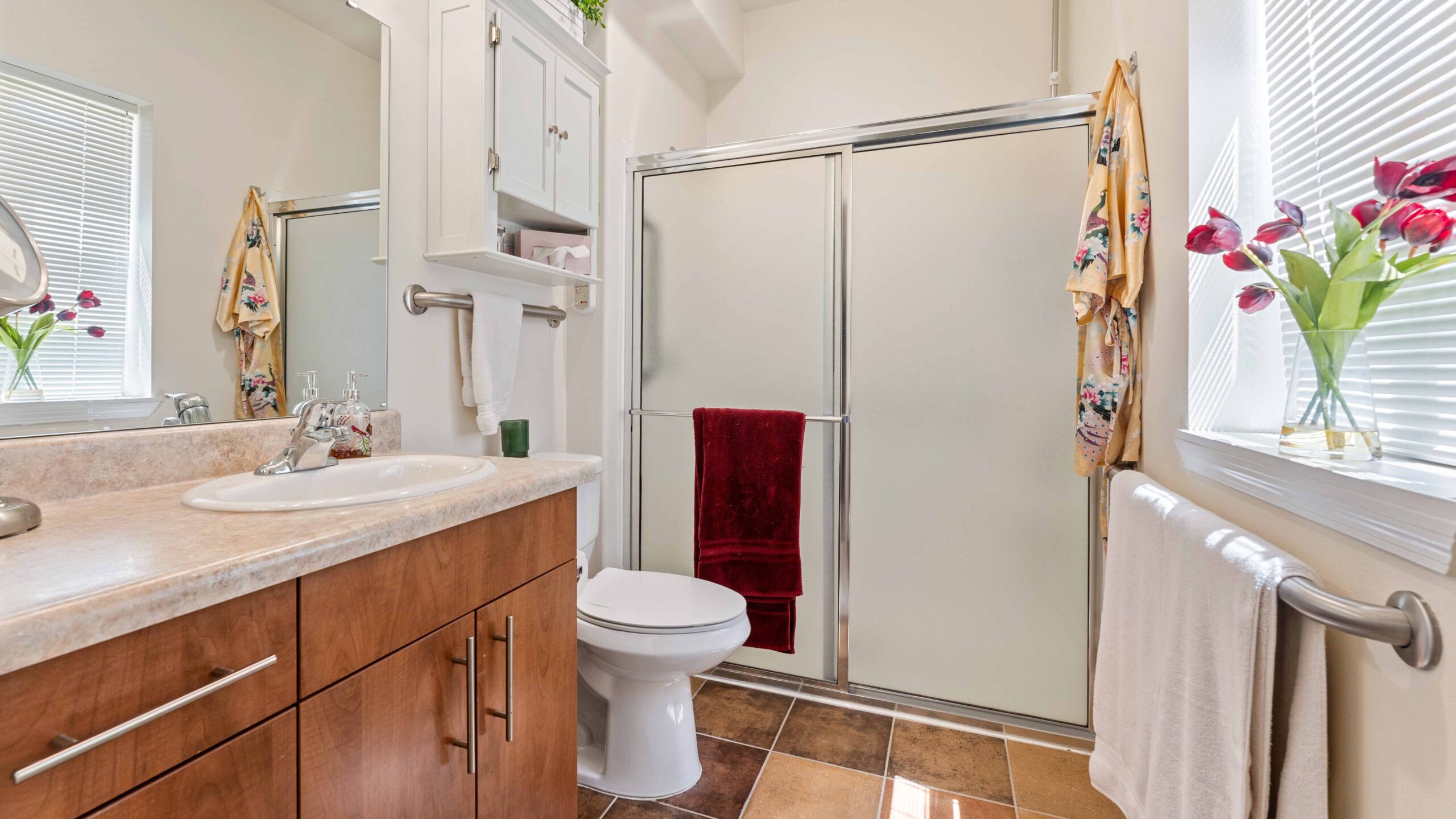 A clean, bright bathroom with a wooden vanity, white sink, toilet, and a glass shower with frosted doors. Red and floral towels hang nearby, and red tulips sit by a window with blinds.
