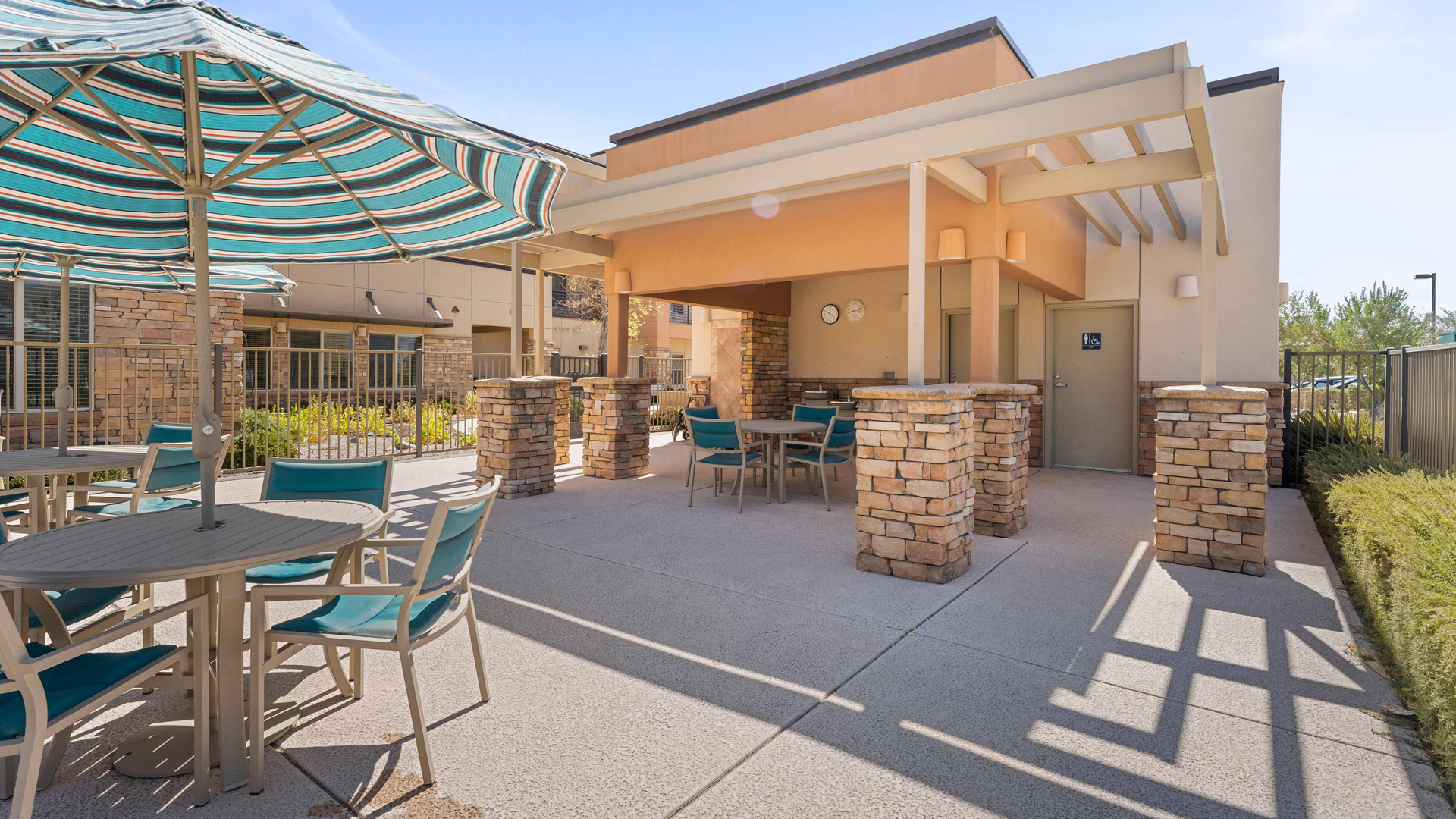 Outdoor patio area with tables and teal chairs, shaded by large striped umbrellas. The patio is attached to a building with stone pillars and a pergola-style roof. Landscaping and a fence are visible in the background.