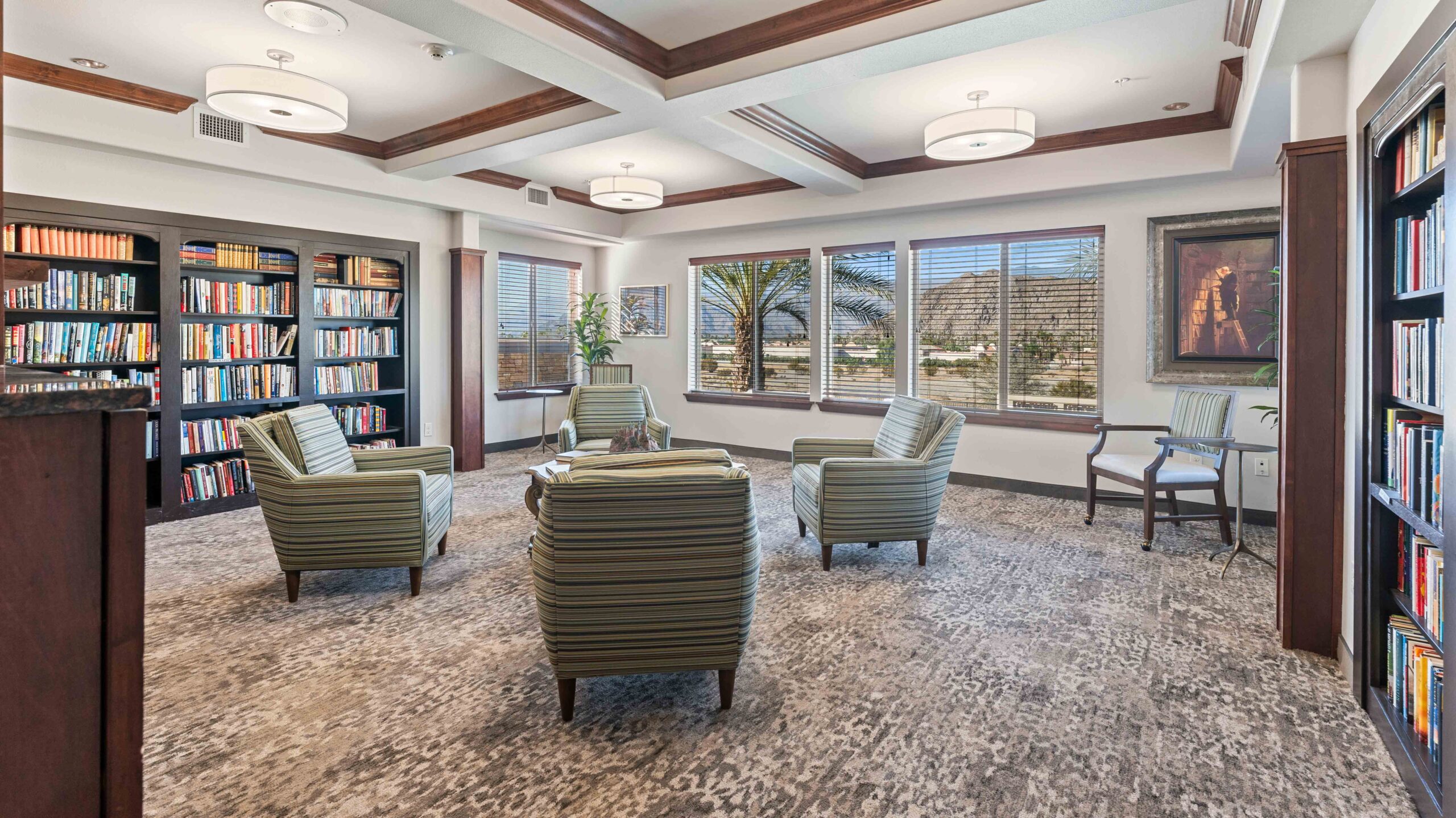 A cozy library room with four striped armchairs arranged in a circle, bookshelves lining the walls, large windows showing a view of palm trees and mountains, and soft overhead lighting.
