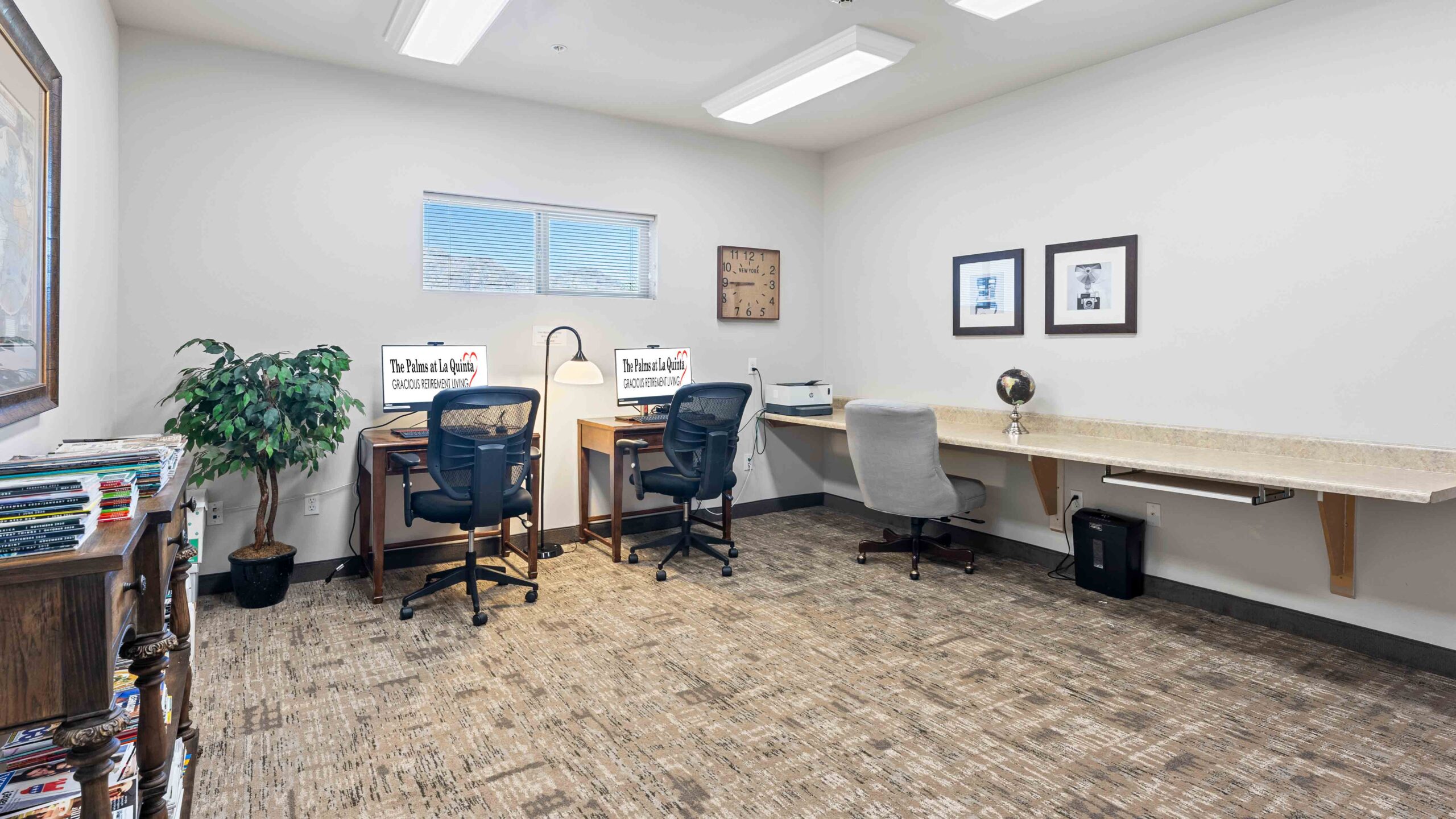 A small office room with three desks, two office chairs, one gray chair, two computer monitors, a printer, framed pictures on the wall, a potted plant, and a shelf with magazines. The room has white walls and a window.