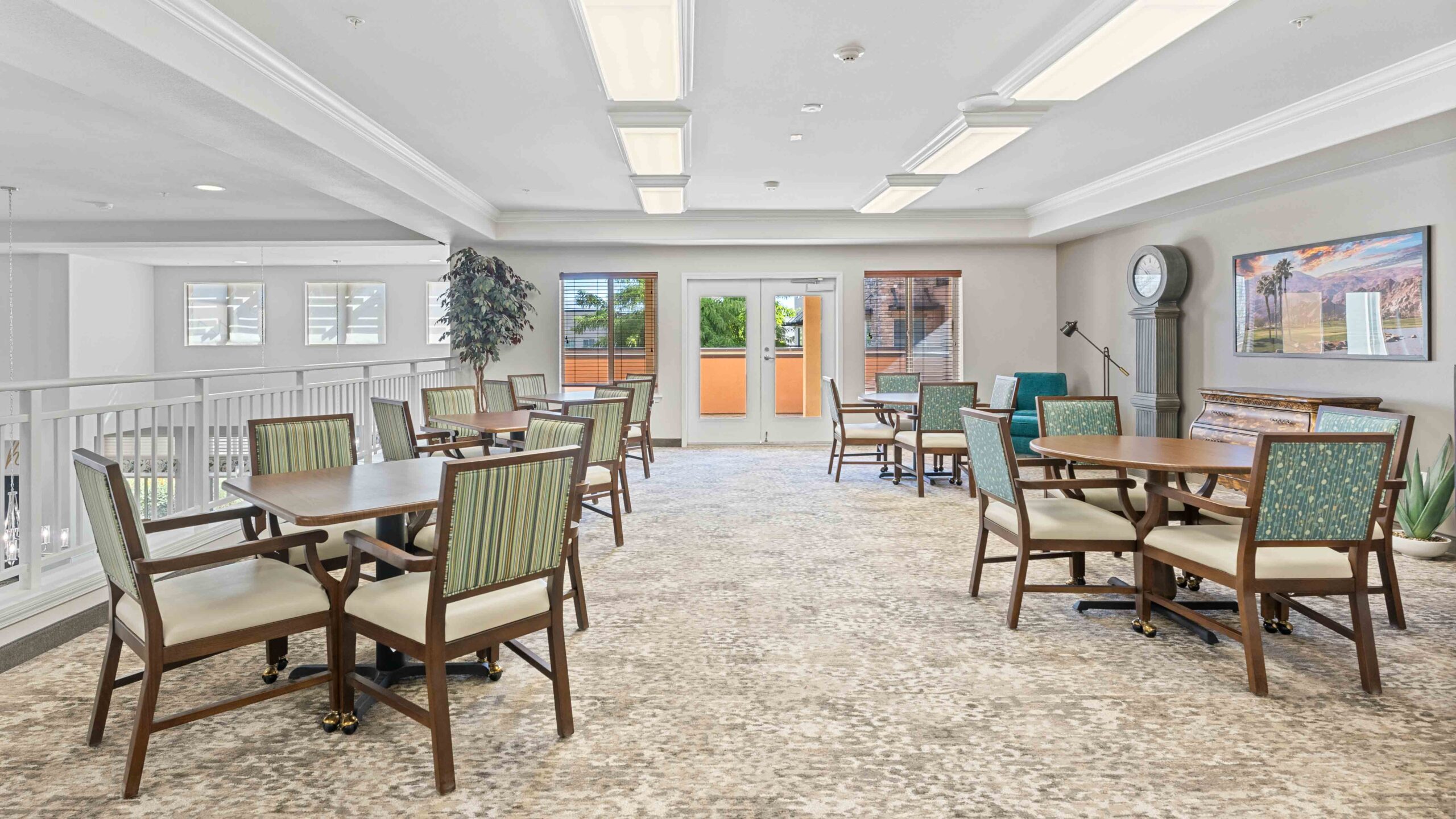 A bright, spacious room with several tables and chairs, large windows, a staircase, a piano, a tall clock, and indoor plants. The decor is neutral with green and wood tones.