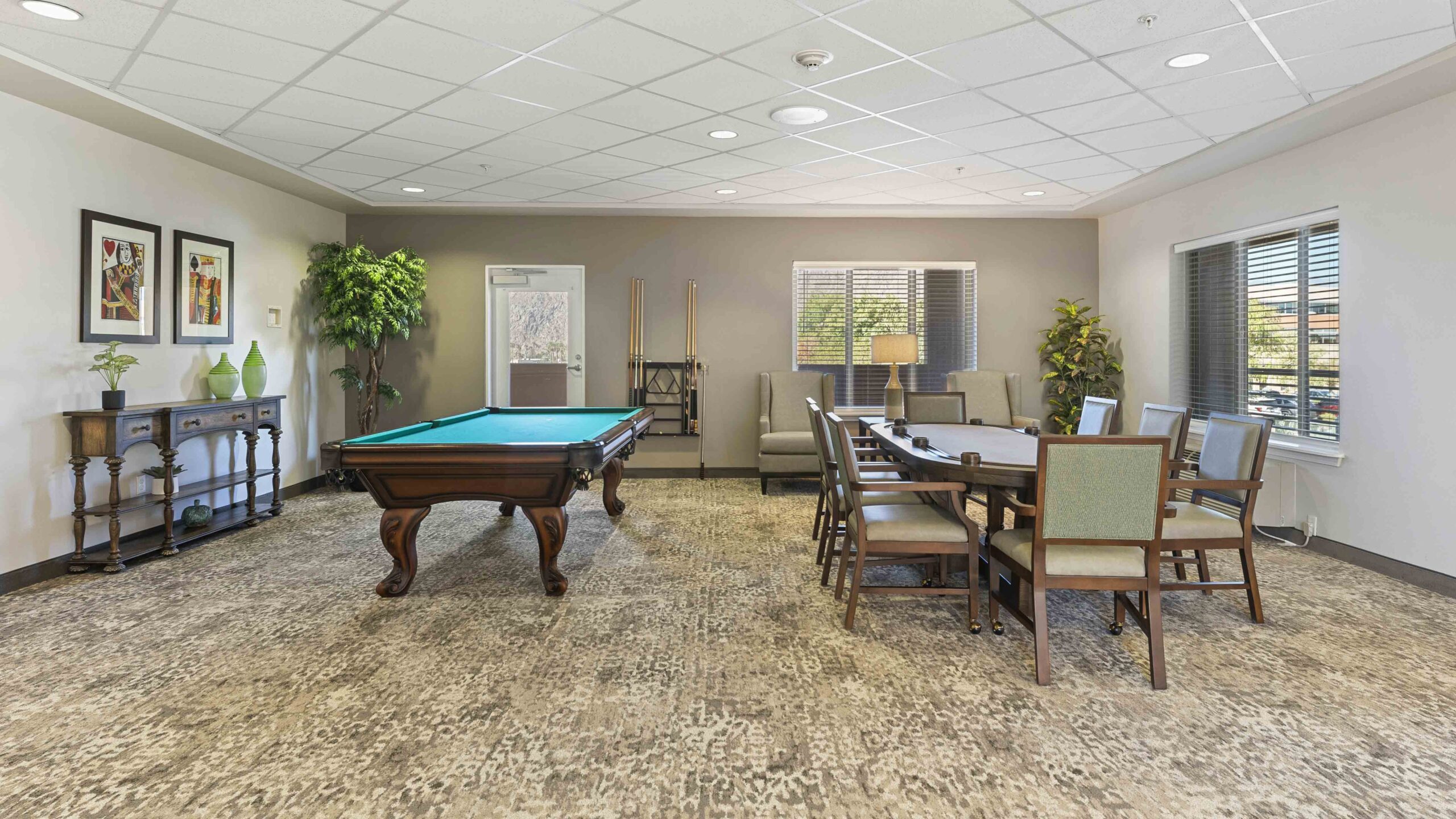 A community game room with a pool table, cue sticks, a seating area with armchairs and a table, plants, framed artwork on the walls, and large windows letting in natural light.
