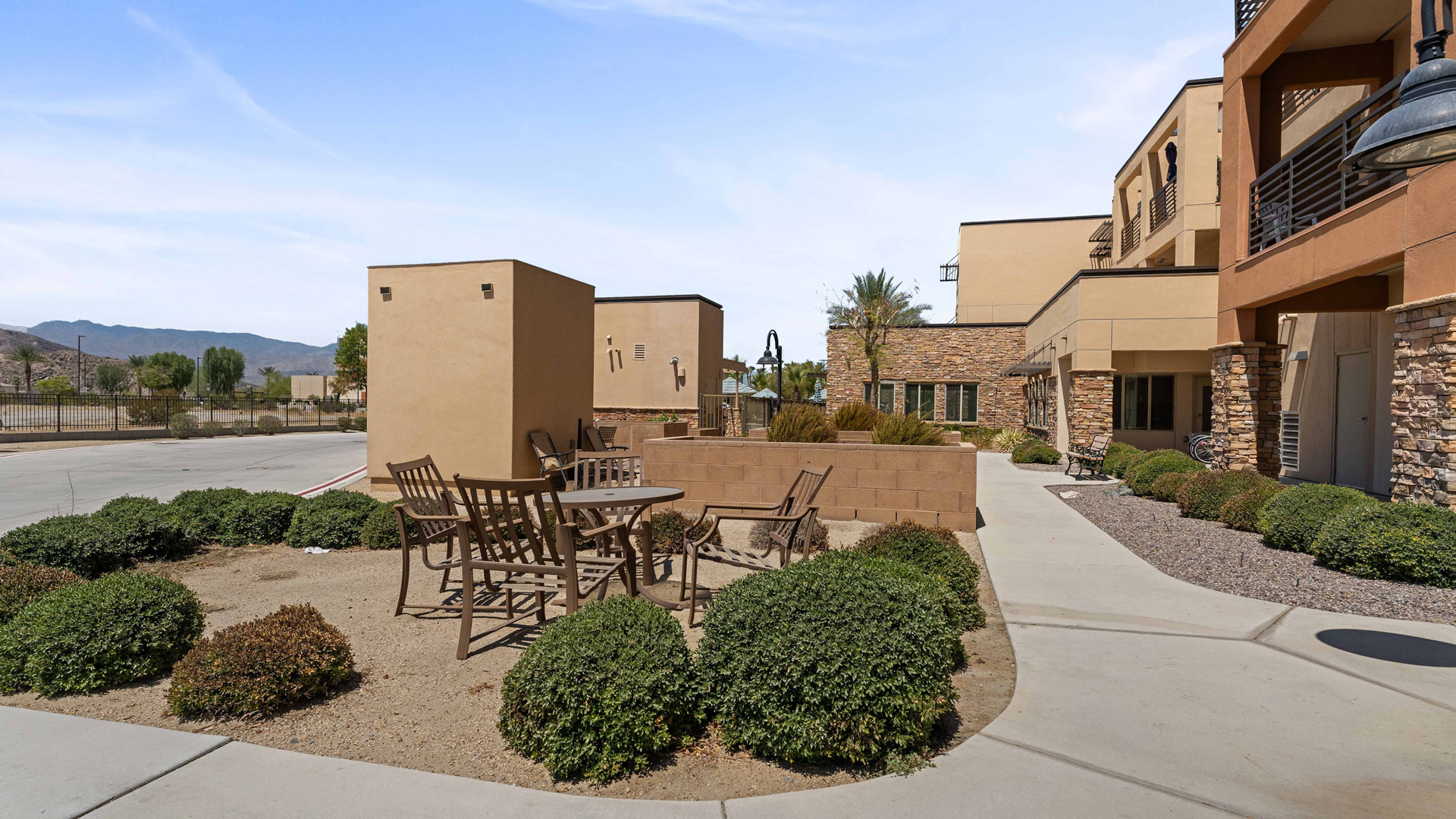 Outdoor patio area with metal tables and chairs surrounded by shrubs, next to a modern beige and stone building under a clear sky; mountains visible in the background.