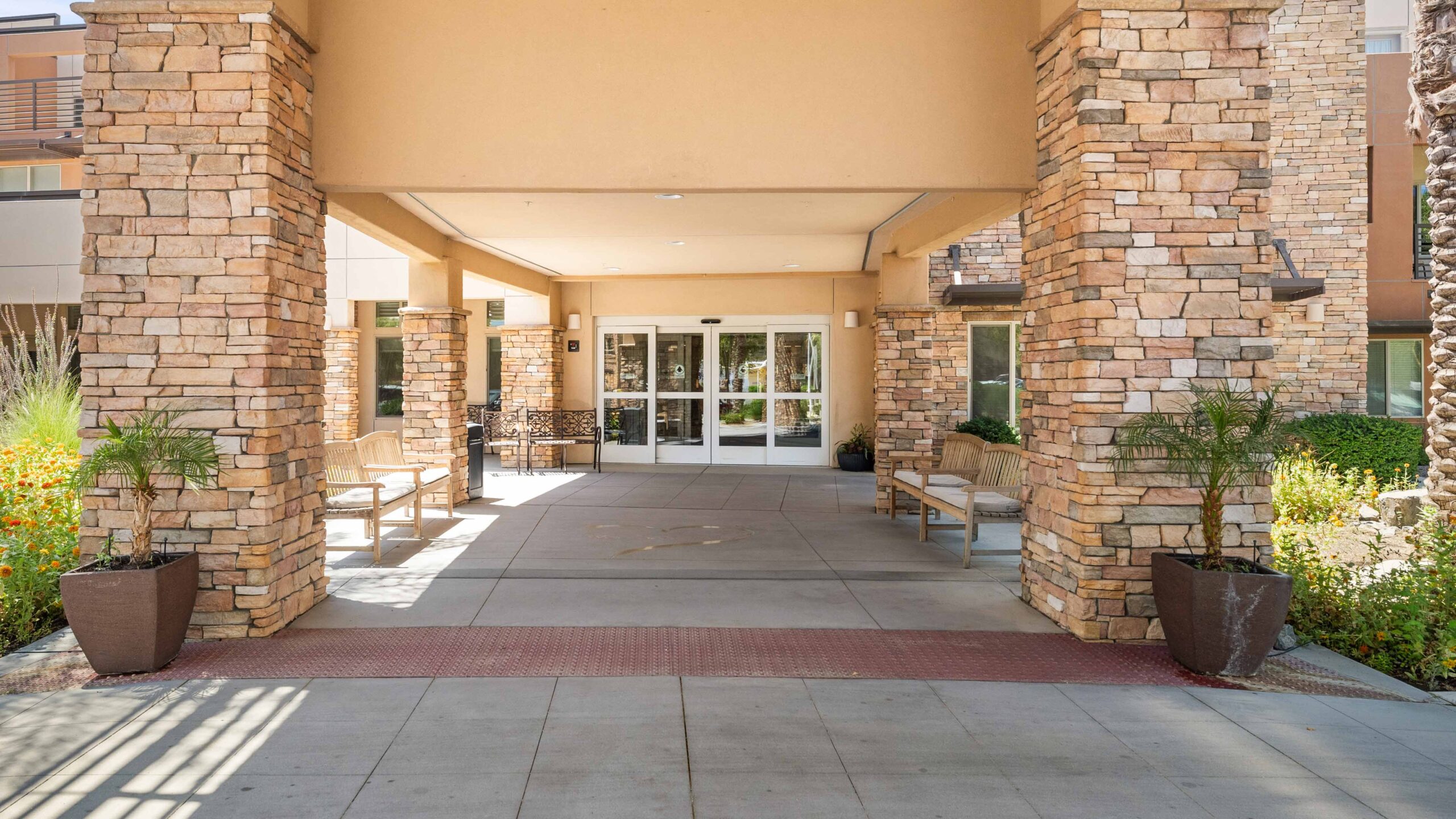 Covered building entrance with stone pillars, glass double doors, benches on both sides, large potted plants, and landscaped greenery along the walkway.