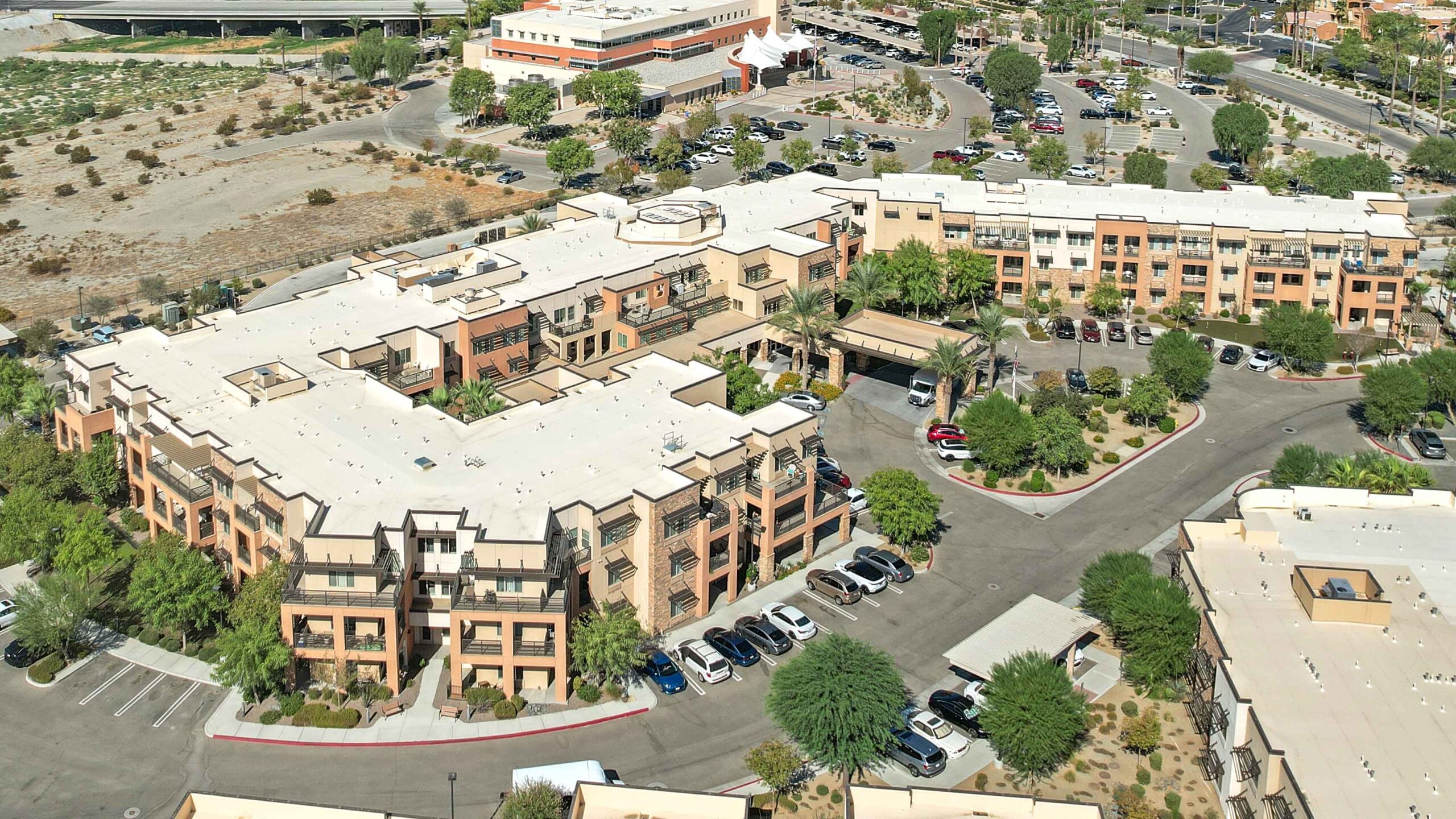 Aerial view of a large, modern, multi-story residential building with a white roof, surrounded by parking lots, landscaped greenery, and nearby roads in a suburban area.