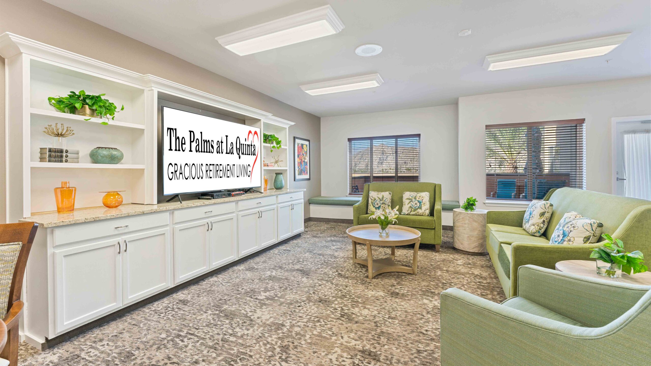 A bright lounge with green sofas, a coffee table, and a large TV displaying “The Palms at La Quinta Gracious Retirement Living.” White shelves with decor line the wall, and sunlight streams through two windows.