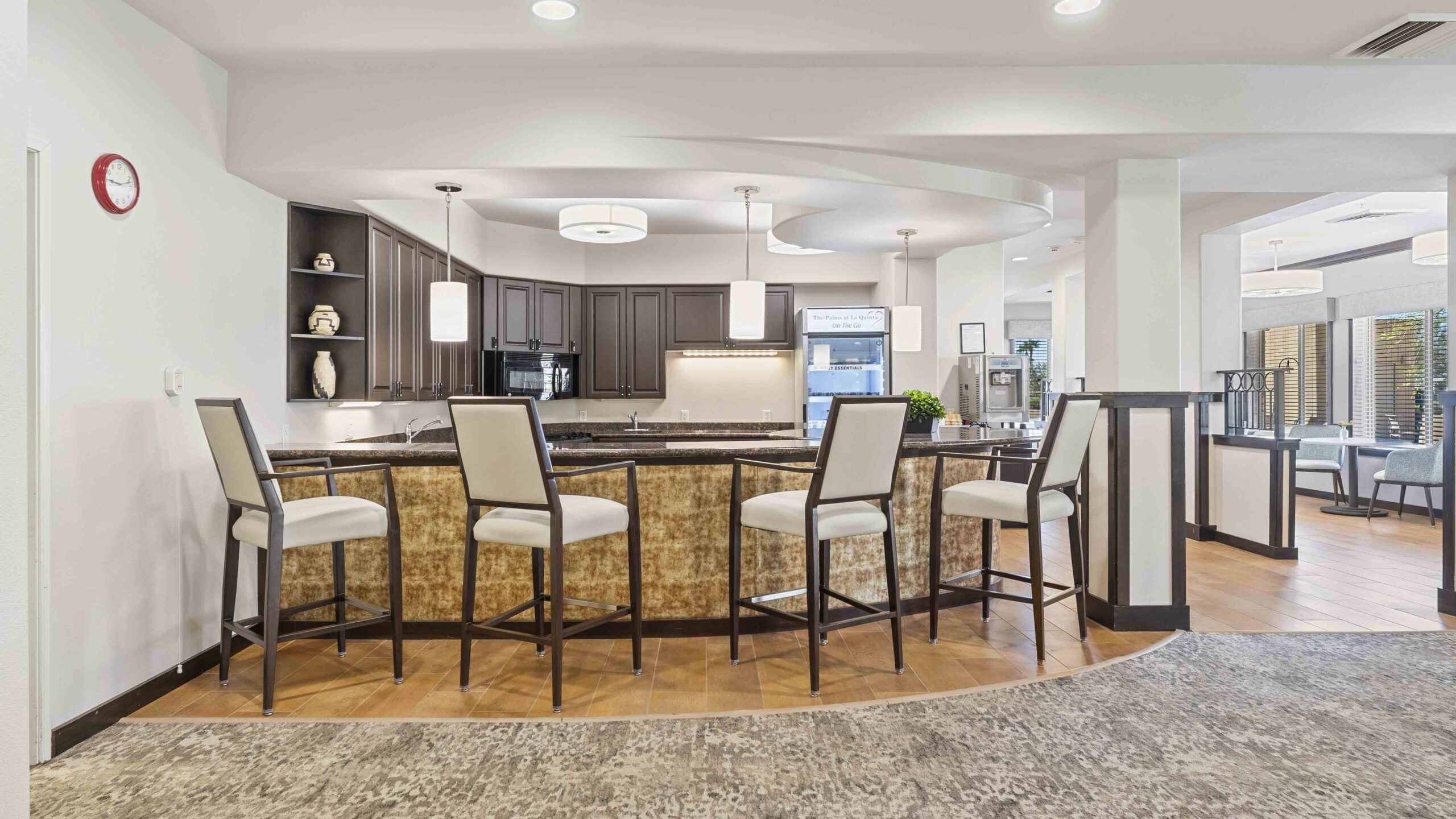 Modern kitchen with a curved bar featuring four high-back chairs, dark wood cabinets, pendant lights, and decorative shelves. The room has light walls, wood flooring, and large windows in the background.