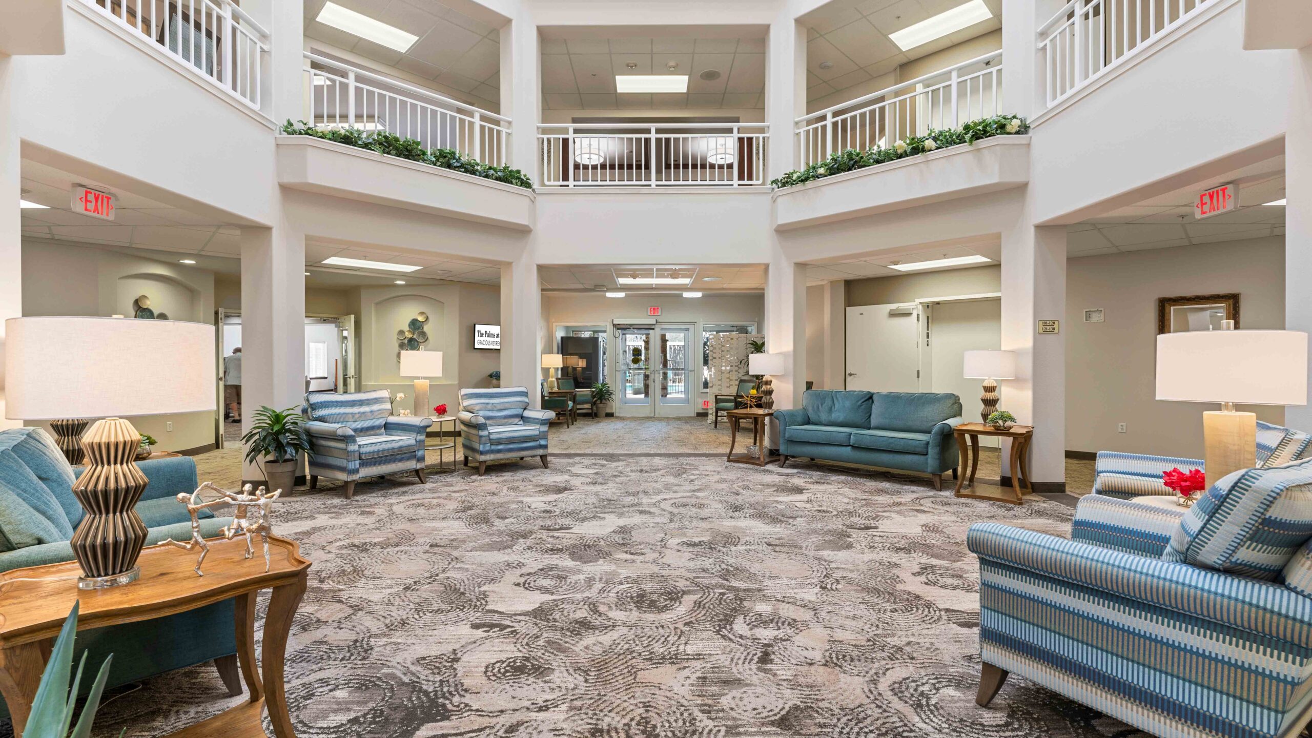 A spacious, bright lobby with patterned carpet, blue sofas and armchairs, side tables with lamps, green plants, and a second-floor balcony with white railings. Double glass doors are visible at the far end.