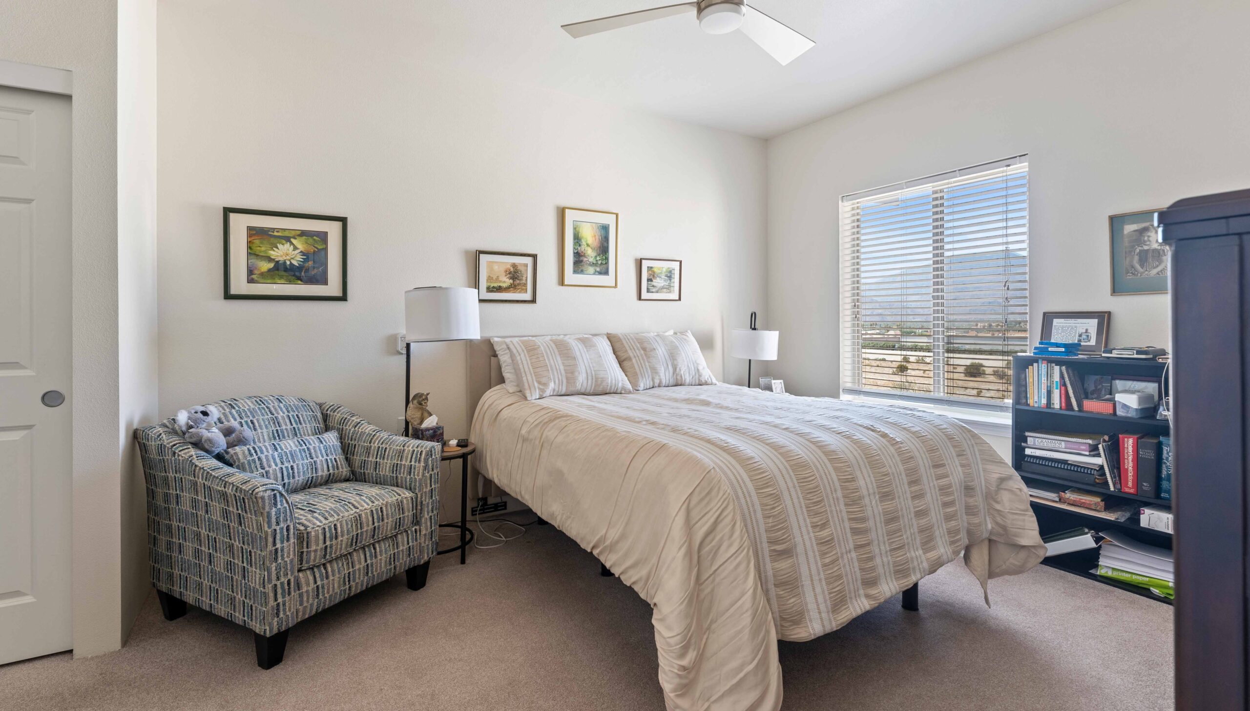 A tidy bedroom with a double bed, a patterned armchair, framed art on the wall, a ceiling fan, a window with blinds, bookshelves, and neutral-colored decor.