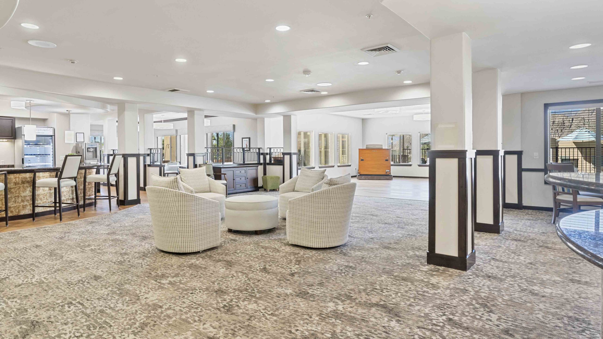 A spacious, modern lounge area with four white armchairs around a circular ottoman, light patterned carpet, bright lighting, and large windows. A bar counter and open seating area are visible in the background.