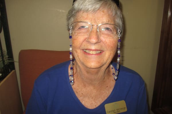 An older woman with short white hair and glasses, wearing a blue shirt and a name tag, is smiling at the camera. She has colorful beaded chains attached to her glasses. She is seated in an indoor setting.