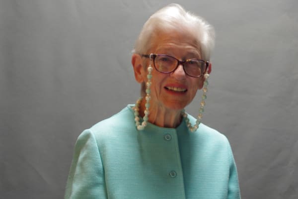 An elderly person with short white hair is wearing glasses with a beaded chain, a light blue outfit, and is smiling at the camera in front of a plain, grey background.