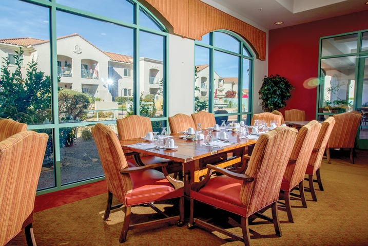 A sunlit dining room with a large wooden table surrounded by upholstered chairs. The table is set with white plates, cups, and saucers. Large windows reveal a view of a courtyard and neighboring buildings with red-tiled roofs. Verdant plants decorate the room.