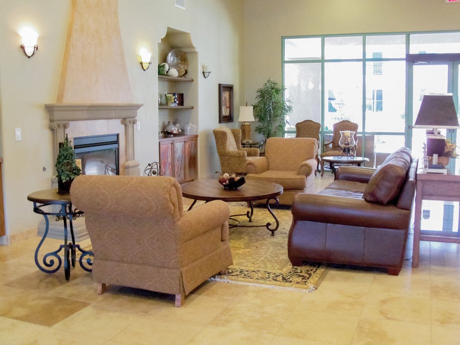 A spacious living room with tiled floors, featuring a mix of armchairs and a leather sofa arranged around a wooden coffee table. There is a fireplace with decorative items on the mantel, built-in shelves, and large windows allowing natural light to fill the space.