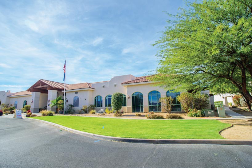 A single-story building with white walls and arched windows stands amidst a landscaped yard with green grass, bushes, and a tree with spreading branches. The sky above is bright blue with thin clouds. An American flag is raised on a flagpole in front of the building.