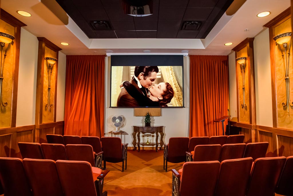 A small, cozy theater room with red upholstered chairs facing a large screen. The screen displays a classic movie scene of a man and a woman embracing. The room's walls are decorated with golden sconces, and long red curtains frame the screen.