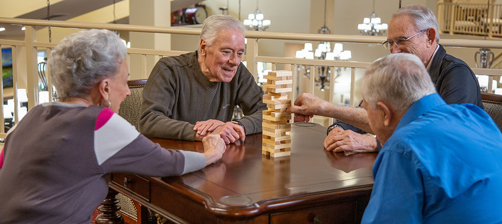 Four elderly individuals are sitting around a wooden table engaged in a game of Jenga. They are smiling and appear to be enjoying the activity in a well-lit, cozy room with chandeliers in the background. One person is carefully removing a block from the tower.