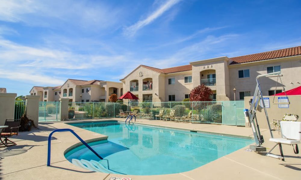 A clear swimming pool is surrounded by a white metal fence. In the background, there is a beige two-story apartment complex with red-tiled roofs. The sky is blue with some clouds, and there are patio tables and chairs with umbrellas near the pool.