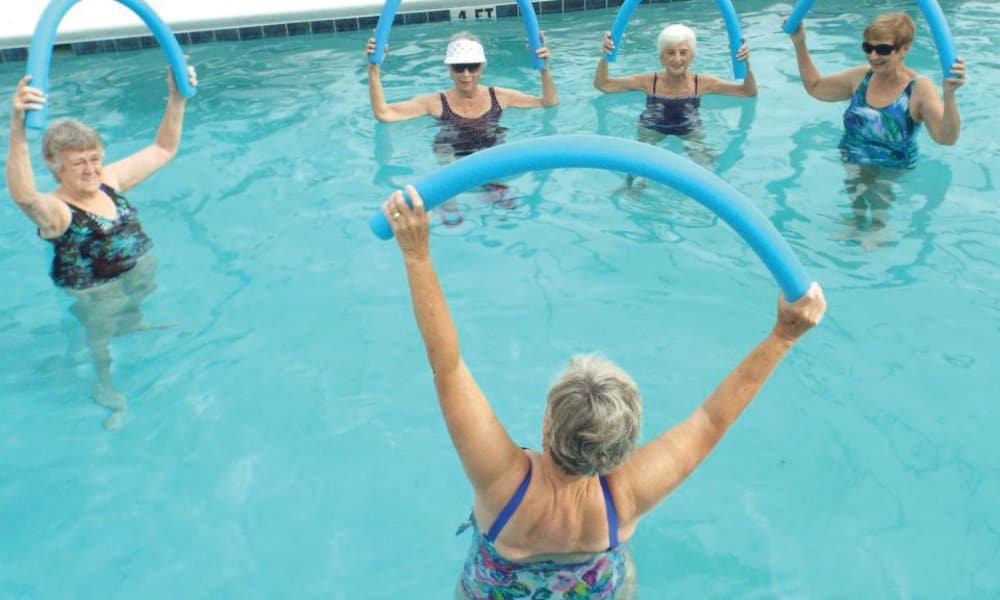 A group of five elderly women in a swimming pool are participating in a water aerobics class. They are holding blue pool noodles above their heads and wearing colorful swimsuits. The instructor stands in the foreground, facing the group with her arms raised, also holding a noodle.