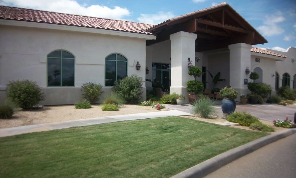 A single-story beige building with a red-tiled roof features large arched windows and a covered entrance supported by pillars. The front is landscaped with green shrubs, small trees, flowering plants, and a neatly manicured lawn under a partly cloudy sky.