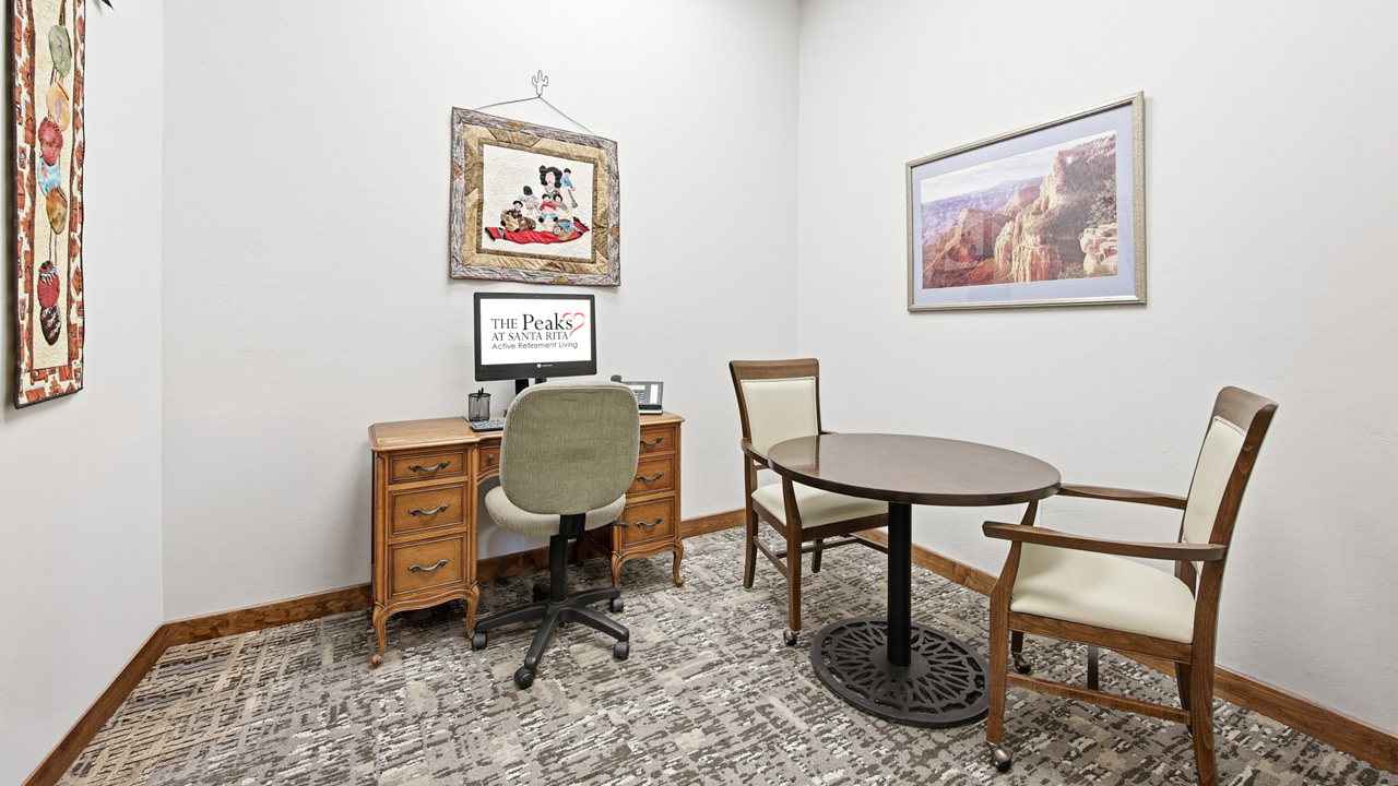 A small office with a wooden desk, green desk chair, computer, and two white-cushioned chairs around a round table. Framed art and a photo hang on white walls above a patterned carpet.