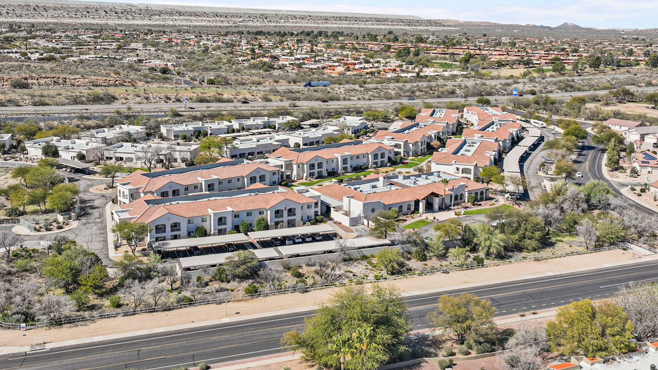 Aerial view of a residential apartment complex with red-tiled roofs, surrounded by desert landscaping and roads, with a highway and more buildings in the background.