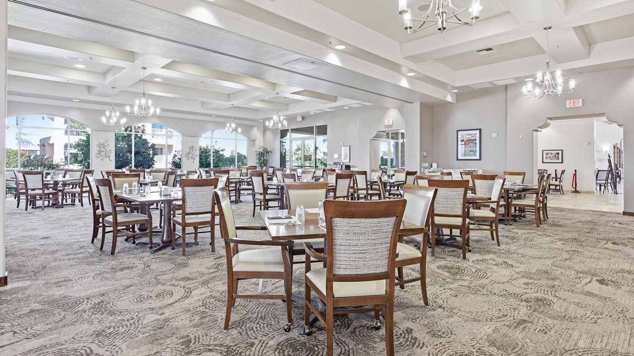 Spacious, well-lit dining room with multiple wooden tables and chairs, neutral decor, chandeliers, large windows, and set for a meal. The room appears clean and organized, with natural light streaming in.