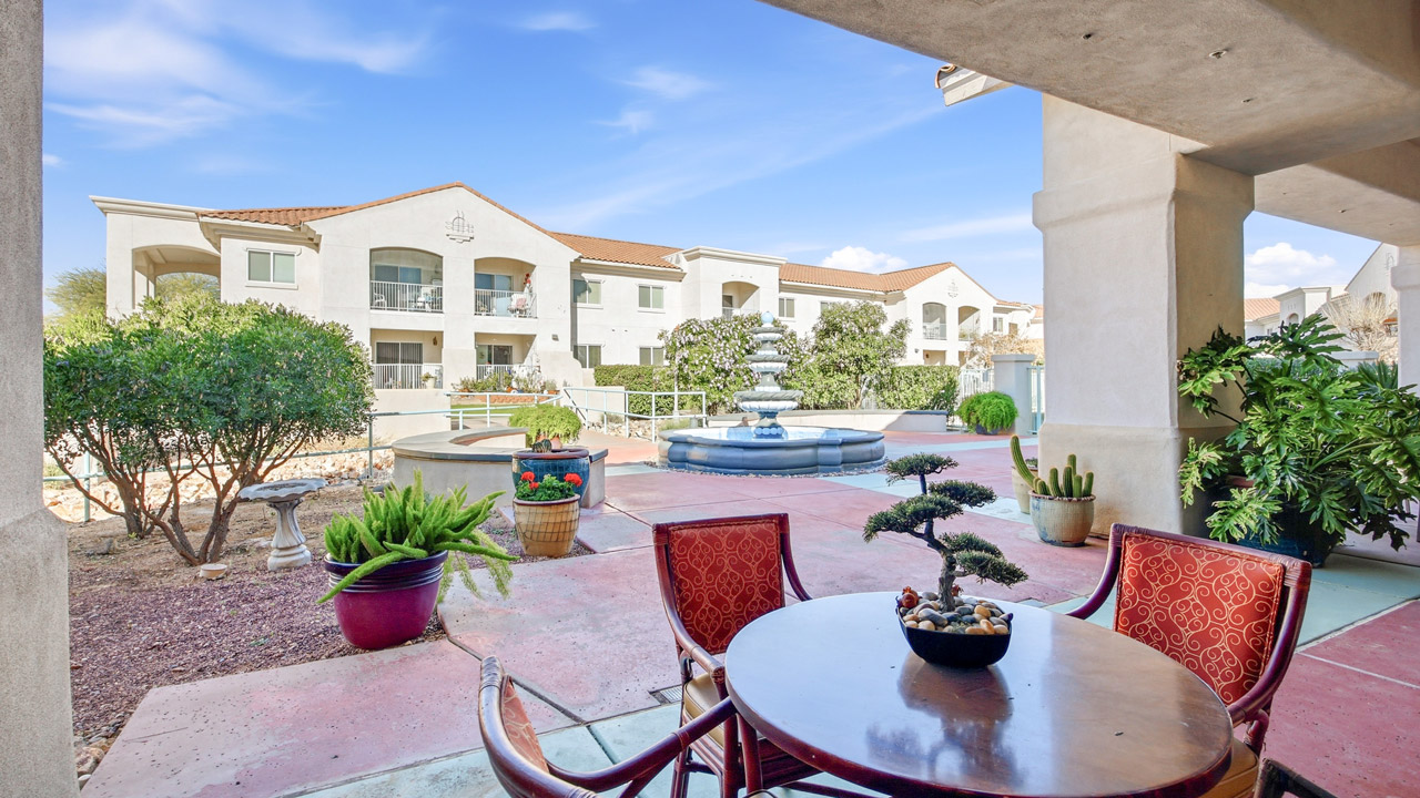 A patio with a round table and red chairs overlooks a courtyard with potted plants, a fountain, and white two-story apartment buildings under a blue sky.
