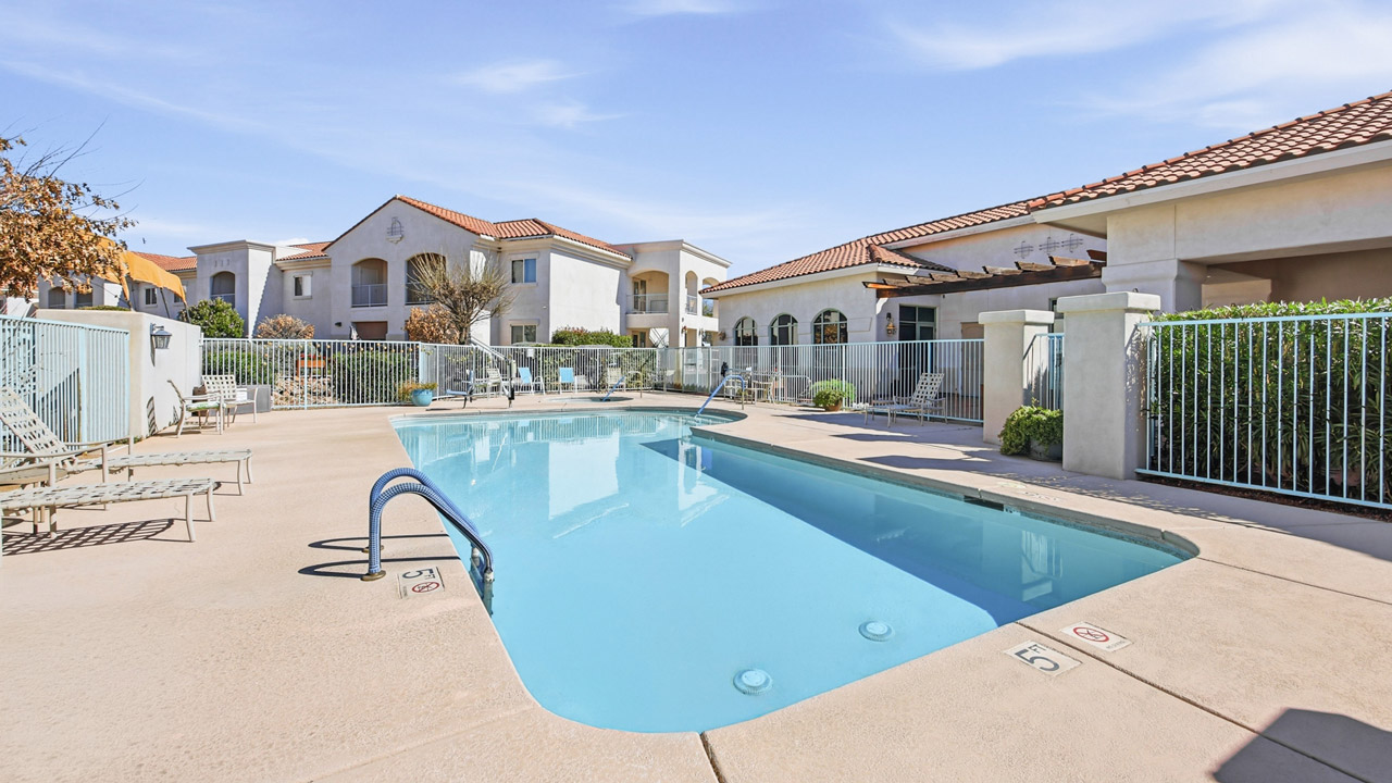 A rectangular outdoor swimming pool surrounded by a fence, lounge chairs, and apartment buildings with red-tiled roofs under a blue sky.