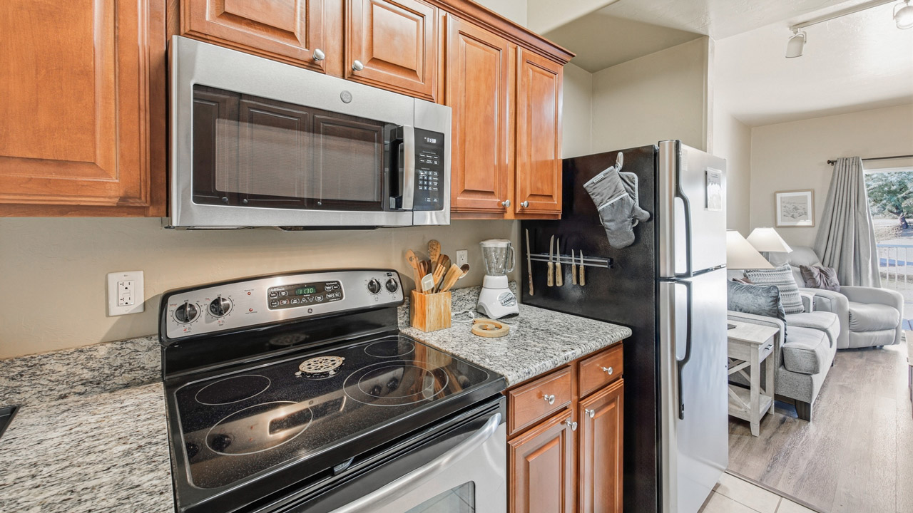 Modern kitchen with wooden cabinets, stainless steel appliances including a stove, microwave, and refrigerator. Granite countertops hold utensils, a blender, and a knife block. Living room with couch is visible in the background.