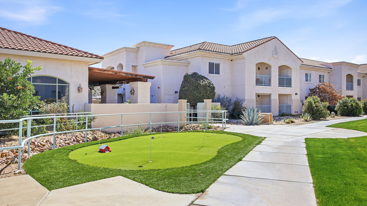 A small putting green with golf clubs and flags sits beside a paved walkway in front of a modern, two-story apartment building with balconies and landscaped gardens under a clear blue sky.