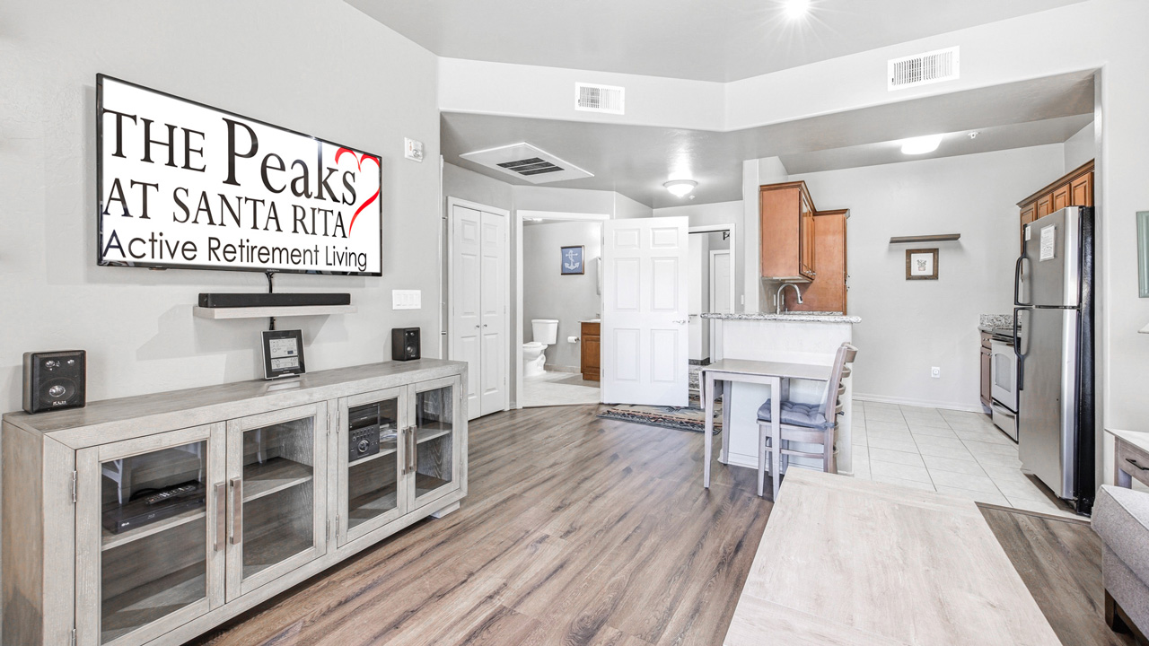 A modern, open-plan apartment with wood flooring, a TV displaying "The Peaks at Santa Rita Active Retirement Living," light wood furniture, a small dining area, and a view into the kitchen and bathroom.