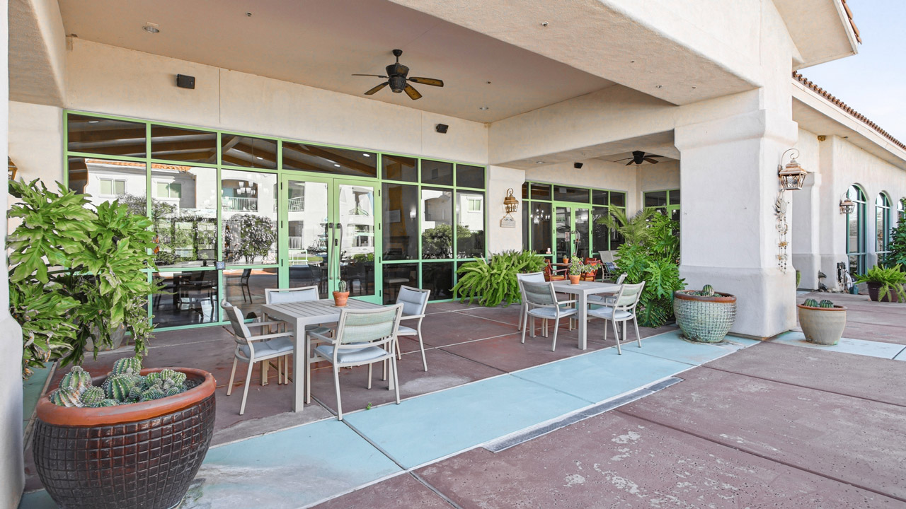 Outdoor patio area with several tables and chairs, potted plants, large windows, ceiling fans, and a covered roof attached to a light-colored building. The atmosphere appears calm and inviting.