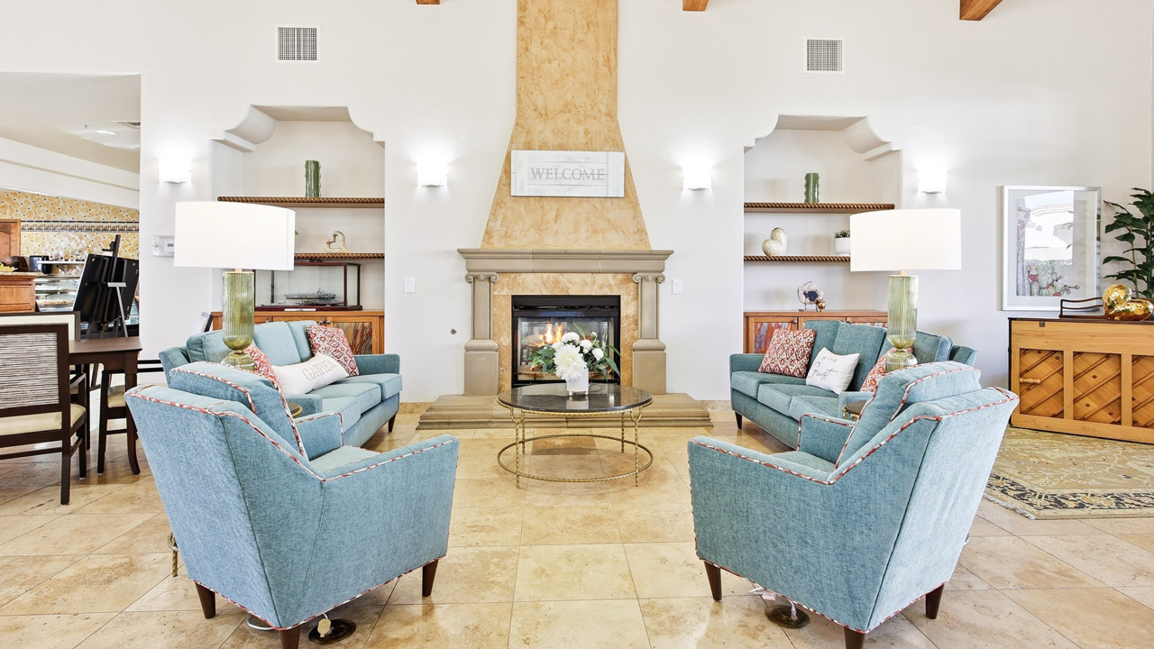 A bright living room with four blue armchairs around a glass coffee table, facing a fireplace. Built-in shelves, table lamps, and decorative pillows add cozy touches. The space has tiled floors and white walls.