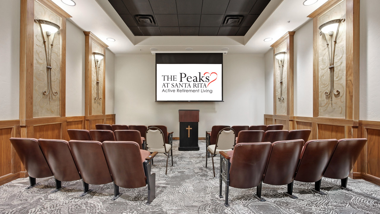 A small chapel-style room with rows of brown chairs facing a podium and a screen displaying “The Peaks at Santa Rita Active Retirement Living.” The room has wood paneling, patterned carpet, and wall-mounted lights.