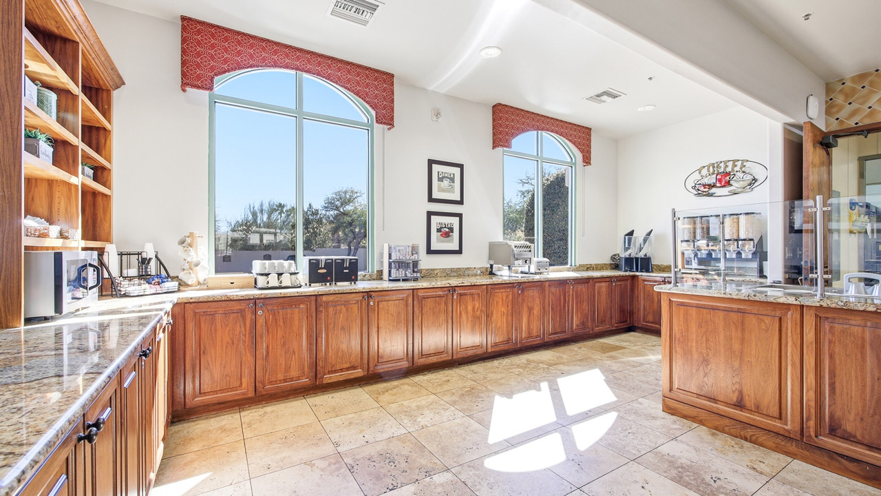 A spacious breakfast area with wooden cabinets, granite countertops, coffee machines, cereal dispensers, and large windows letting in natural light. The floor is tiled, and framed art hangs on the white walls.