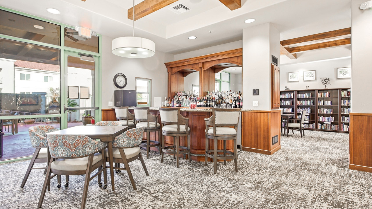 A bright lounge with a round table and chairs, a wooden bar stocked with bottles, shelves of books in the background, and large windows letting in natural light.