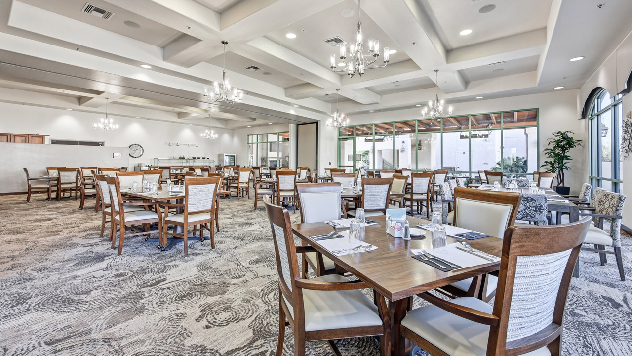A spacious, well-lit dining room with wooden tables and chairs neatly arranged, set with placemats and silverware. Large windows let in natural light, and modern chandeliers hang from the ceiling.