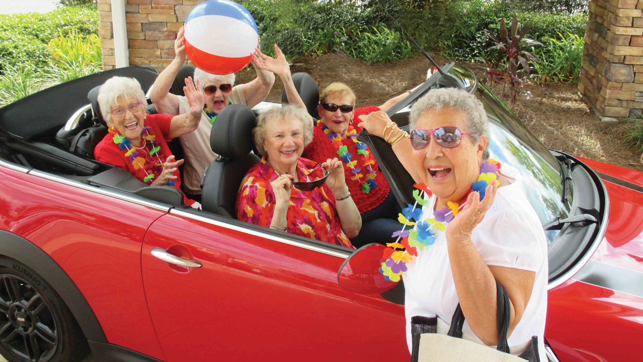 A group of cheerful older women in colorful leis and sunglasses smile and wave while sitting in and around a red convertible. One woman outside holds a beach bag, and another holds a beach ball. The scene looks festive and fun.