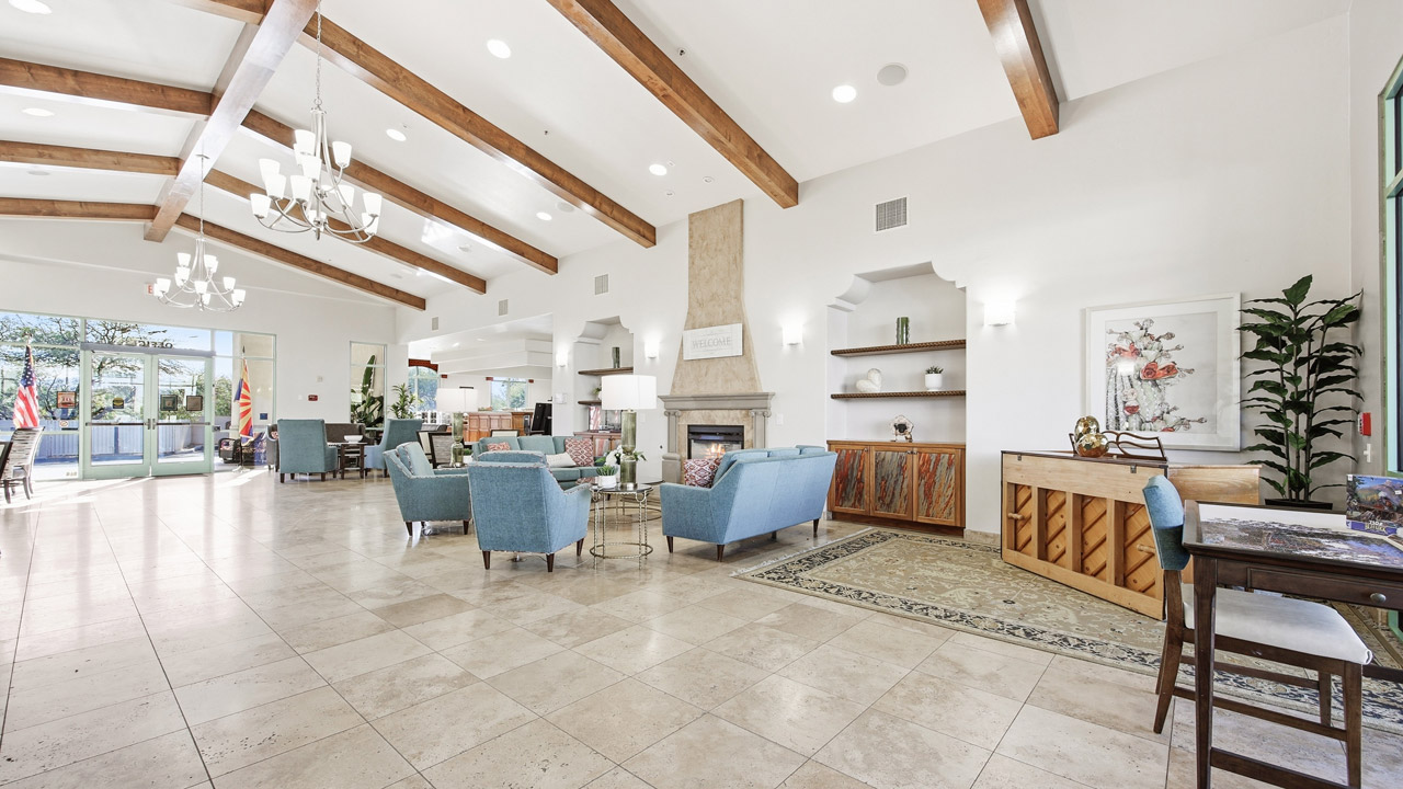 A spacious, bright lobby with beige tiled floor, blue armchairs around a fireplace, wooden ceiling beams, chandeliers, and large windows letting in natural light. There are plants and American flags near the entrance.