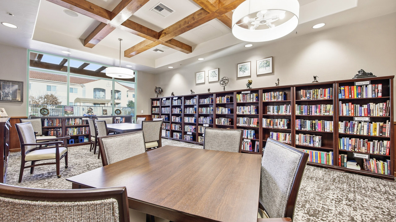 A modern library room with wooden tables and chairs, bookshelves filled with books along the walls, large ceiling lights, and a window letting in natural light. Framed art and small sculptures decorate the space.