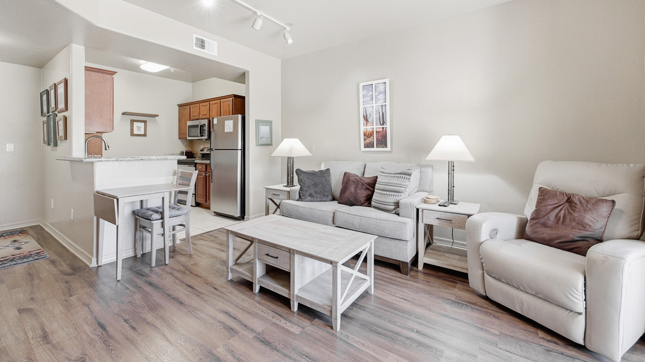 A modern living room with wood flooring, a gray sofa, a beige recliner, a coffee table, two side tables with lamps, and a small dining table near an open kitchen with stainless steel appliances.