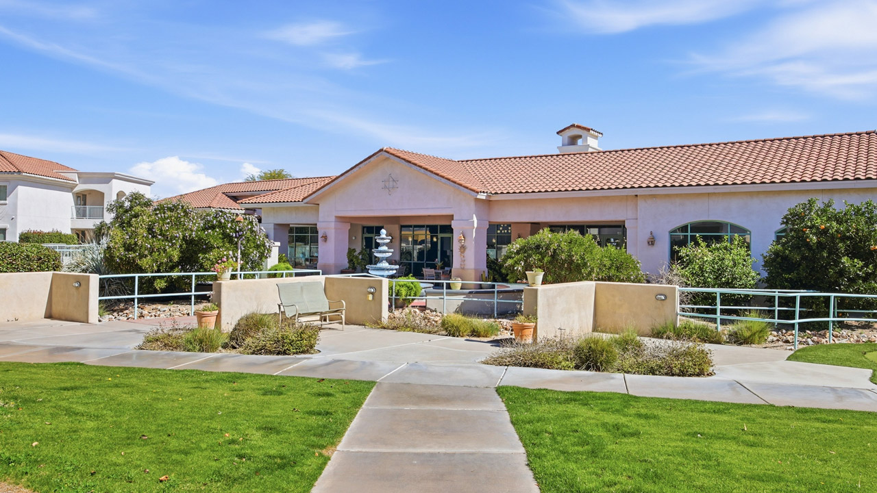 Single-story building with a red tile roof, landscaped gardens, a central walkway, benches, potted plants, and a fountain in front. The sky is blue with some clouds.