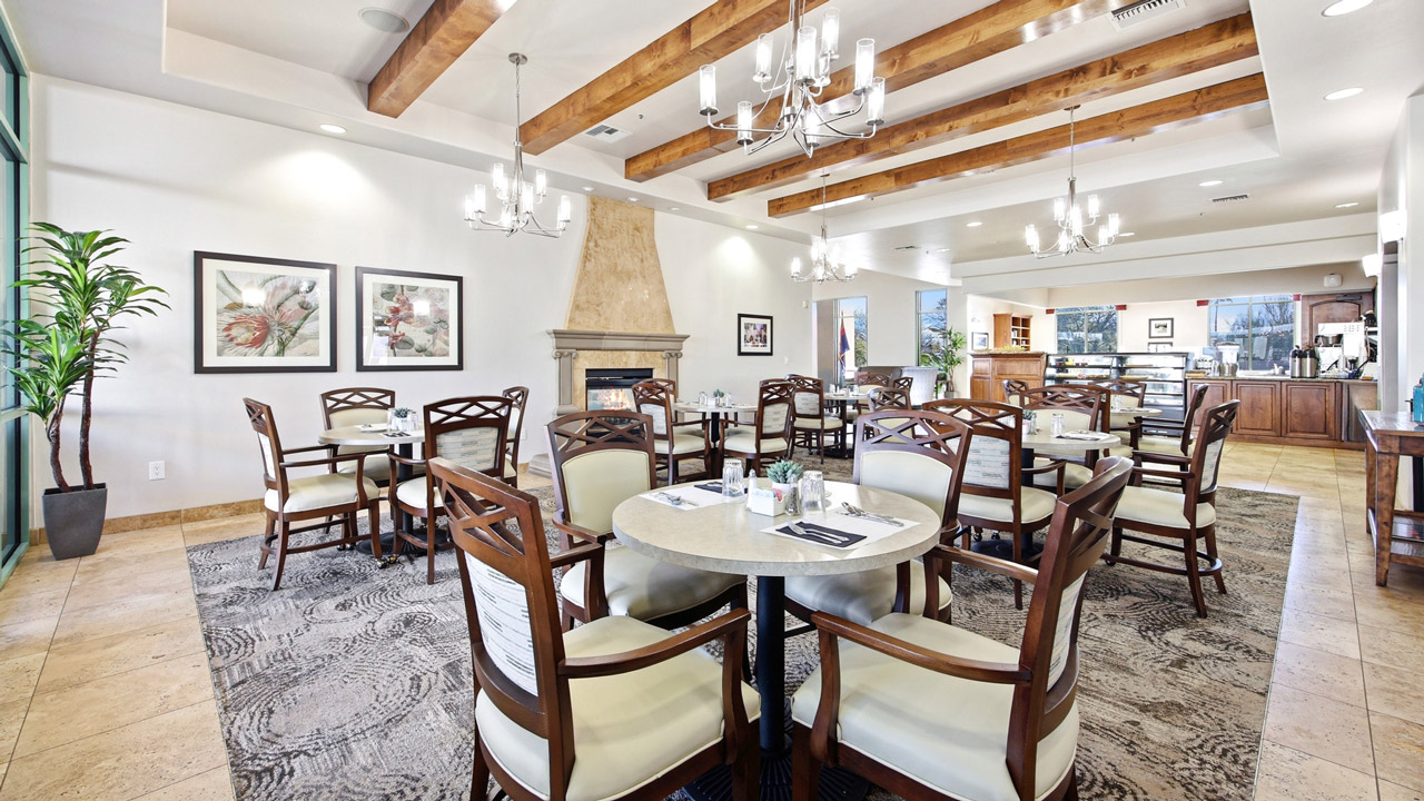 A bright, elegant dining room with wooden ceiling beams, chandeliers, several round tables with cushioned chairs, a stone fireplace, framed art, large windows, and a buffet area in the back.