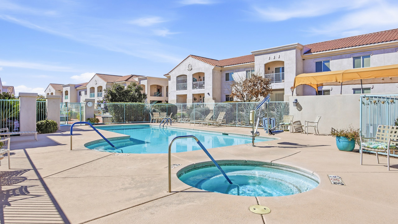 Outdoor swimming pool area with a hot tub, surrounded by a concrete deck, lounge chairs, and fencing, set against a backdrop of modern apartment buildings under a clear blue sky.