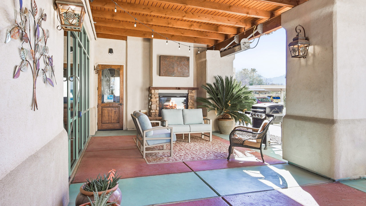 Covered patio with wood ceiling, outdoor fireplace, seating area with cushioned chairs and a table, a grill, potted plants, art on the walls, and string lights, with sunlight streaming in from the side.