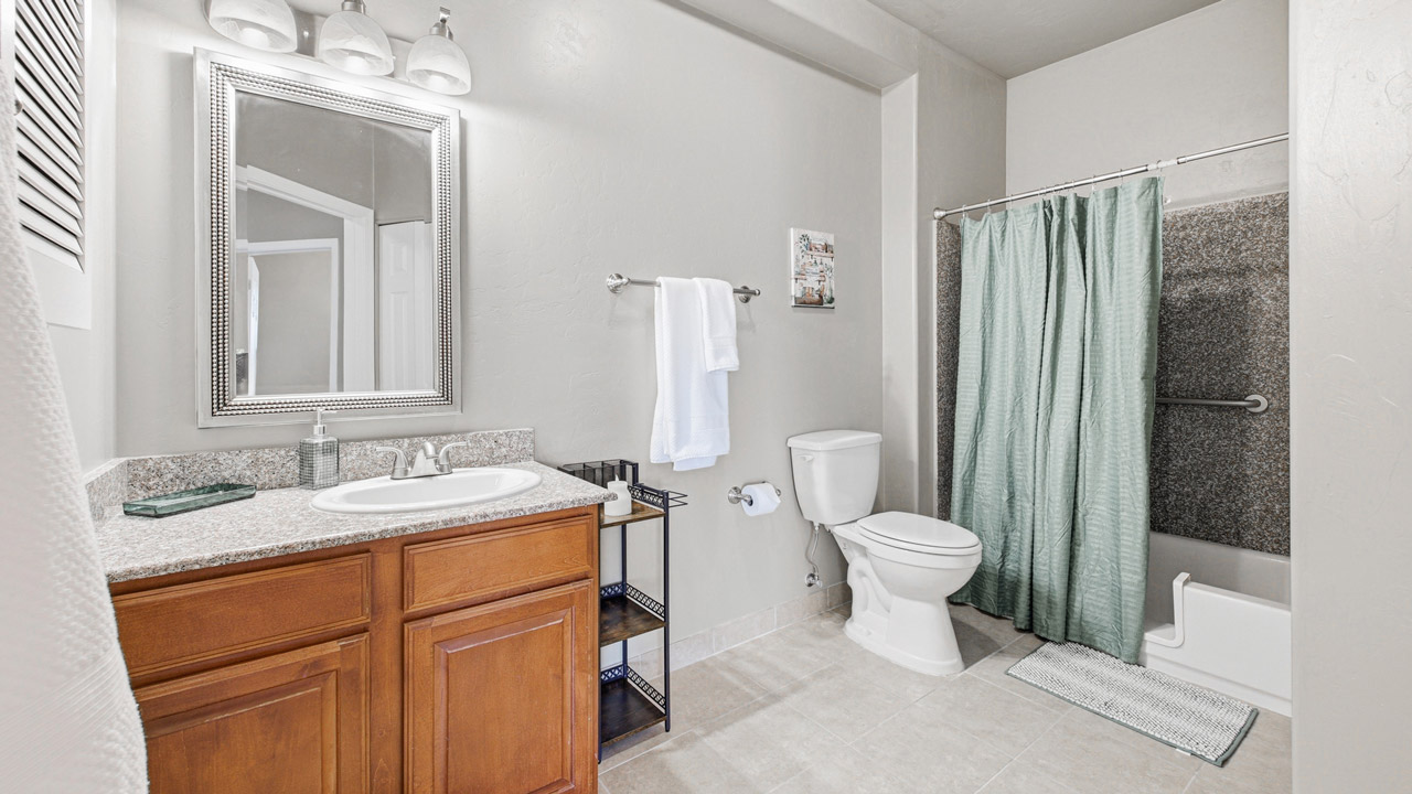 A clean, modern bathroom with light walls, a wooden vanity with a sink and mirror, a toilet, towel rack, and a shower with a green curtain. The floor is tiled and bath essentials are neatly arranged.